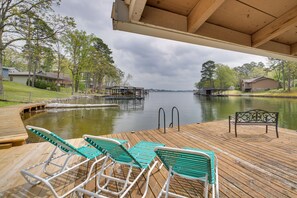 Une terrasse en bois avec des chaises longues et un banc, d’où l’on a vue sur un lac paisible, le tout entouré d’arbres et de maisons.