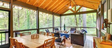 Una terraza techada con mesa y sillas de madera, mobiliario de mimbre y ventanas amplias con vistas al bosque.