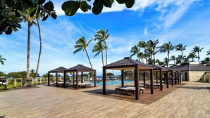 Terraza de madera con mobiliario exterior y piscina, rodeada de palmeras y un cielo despejado.