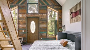 A bedroom with a wooden ceiling, a bed with floral bedding, a ladder, and a door with a stained glass window. - Colorful Cabin w/ Teepee, Fire Pits & Mtn Views! (Marble)