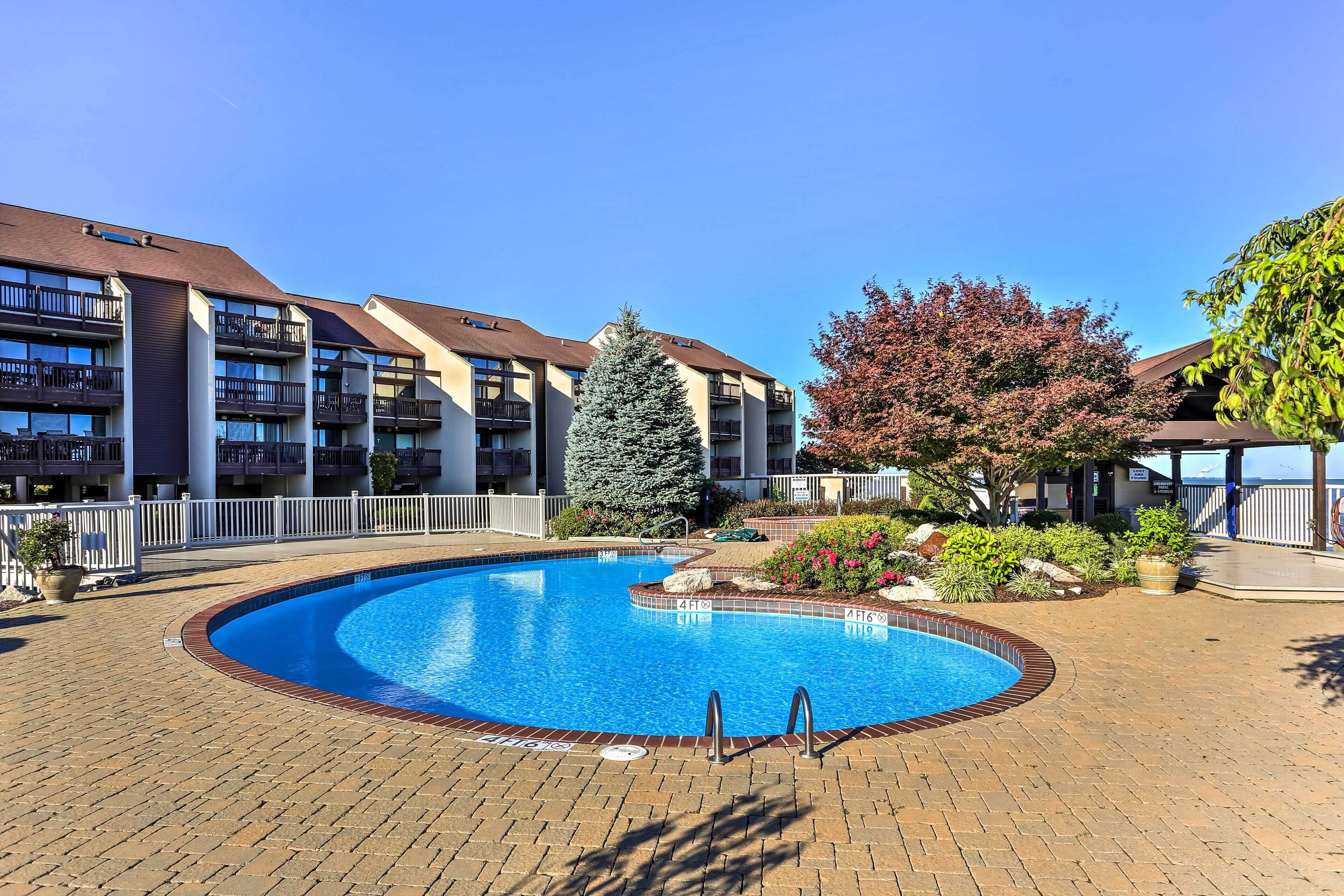 A swimming pool surrounded by a brick patio, with a white fence and a large evergreen tree.