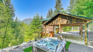 Una baita di legno con vasca idromassaggio e patio coperto, immersa tra alberi e con una montagna sullo sfondo.
