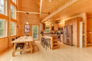 Une cabane en bois spacieuse, dotée d’une grande table à manger, de tabourets de bar, d’une cuisine équipée d’appareils électroménagers en acier inoxydable et de grandes fenêtres.