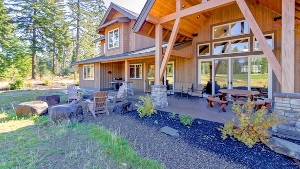 Una cabaña de troncos moderna con un amplio patio, mobiliario exterior y vistas al bosque circundante.