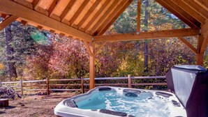 Un bain à remous installé sous une pergola en bois, avec en arrière-plan un paysage naturel composé d’arbres et une clôture en bois.