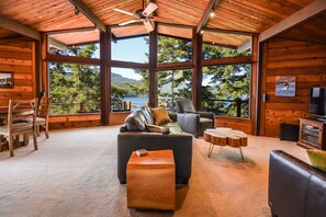 Uma sala de estar ampla com uma grande janela com vista para um lago, um ventilador de teto e uma área de estar aconchegante com um sofá e poltronas.