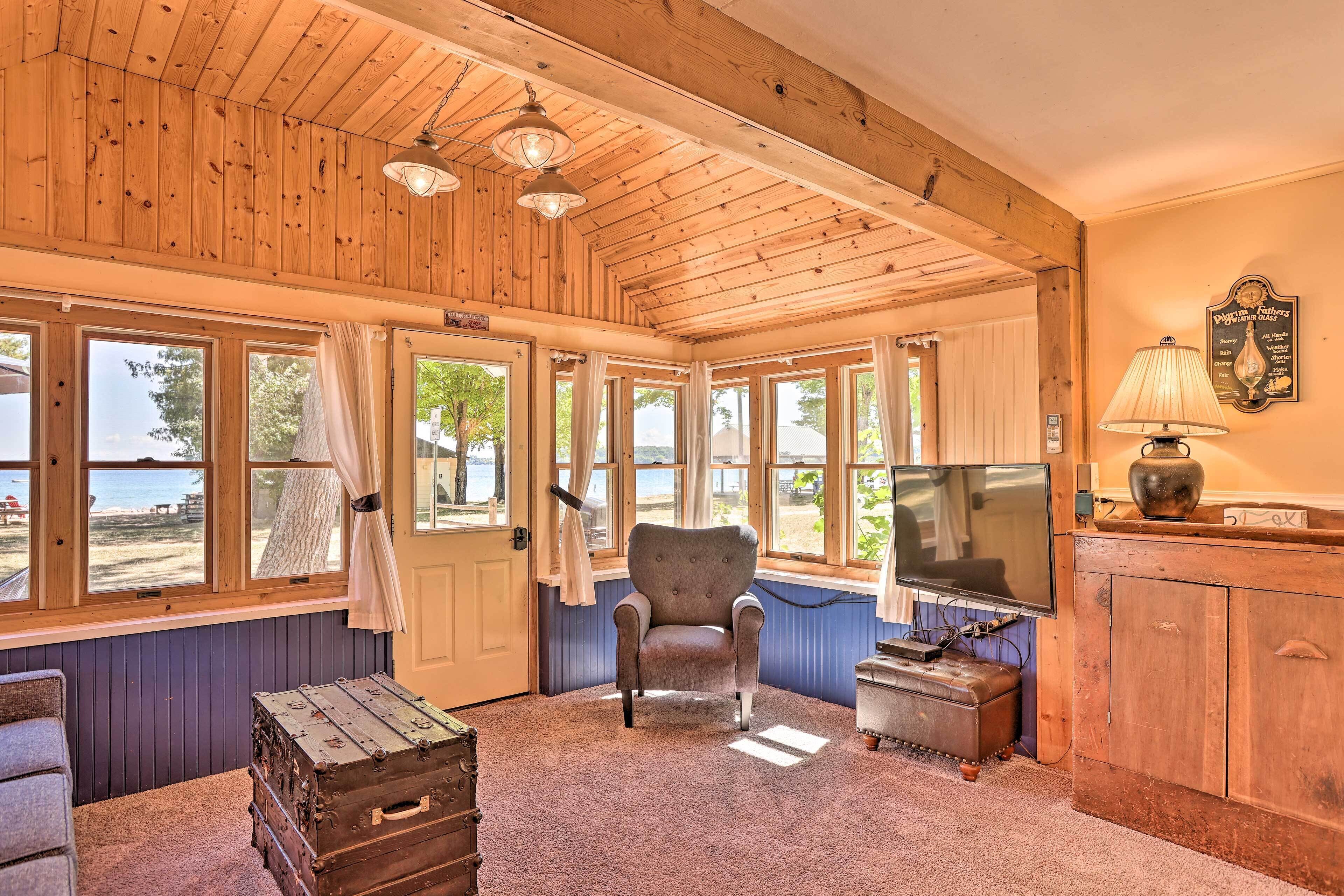 Ein gemütliches Zimmer mit Holzbalkendecke, großen Fenstern, einem bequemen Sessel, einem Fernseher und einer Vintage-Lampe.