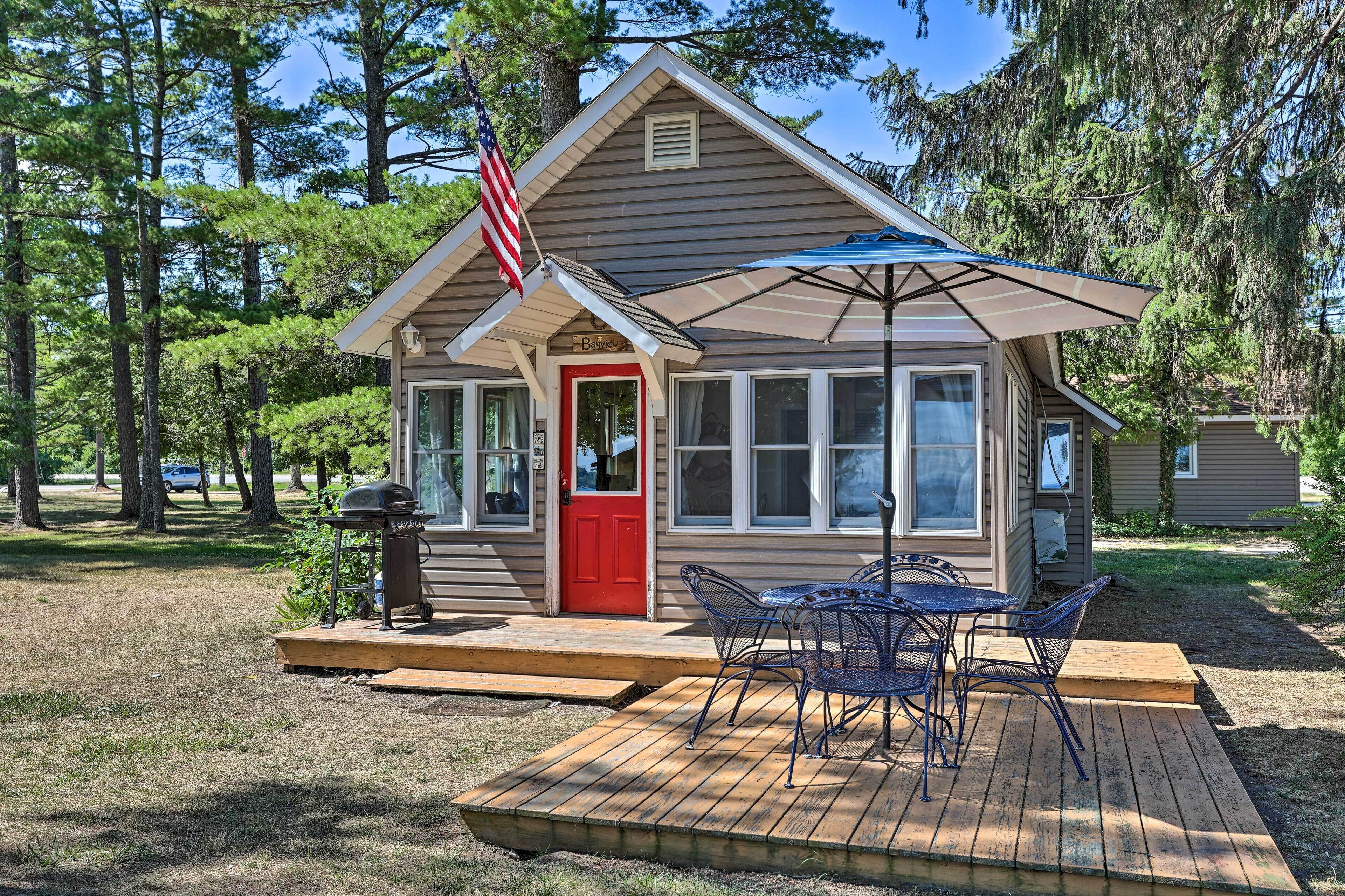 A small cabin with a red door, a deck with outdoor furniture, and an American flag.