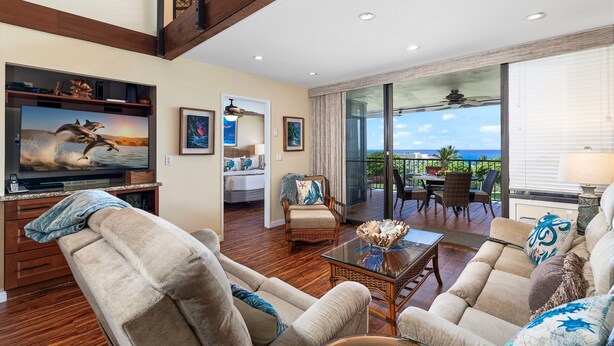 A spacious living room with a large flat-screen TV, comfortable seating, a dining area, and a balcony with a view.