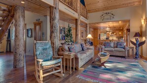 A spacious living room with a high ceiling, wooden beams, a large wooden column, a rocking chair, a beige sofa, a colorful rug, and a Christmas tree. - Tippy Canoe By Tahoe Getaways - Hot Tub & Sauna + EV Charger (Tahoma)