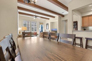 Un coin repas avec une table et des chaises en bois, un ventilateur de plafond et une cuisine visible à travers une porte ouverte.