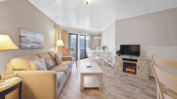 A modern living room with a sofa, coffee table, TV, fireplace, and dining area. - 107 Pointes North Inn | Downtown Charlevoix Gem | Indoor/Outdoor Pool (Charlevoix)