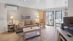 A modern living room with a sofa, coffee table, and TV. There is a window with curtains, a lamp, and an air conditioner on the wall. - 107 Pointes North Inn | Downtown Charlevoix Gem | Indoor/Outdoor Pool (Charlevoix)