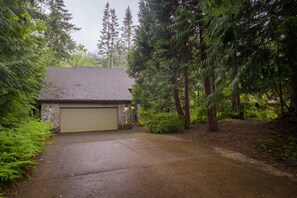 Une maison avec une façade en pierre, un garage et une allée bordée d’arbres denses.