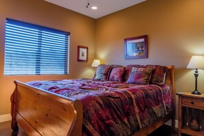 A bedroom with a wooden bed, a window with blinds, a bedside table with a lamp, and two framed pictures on the wall.