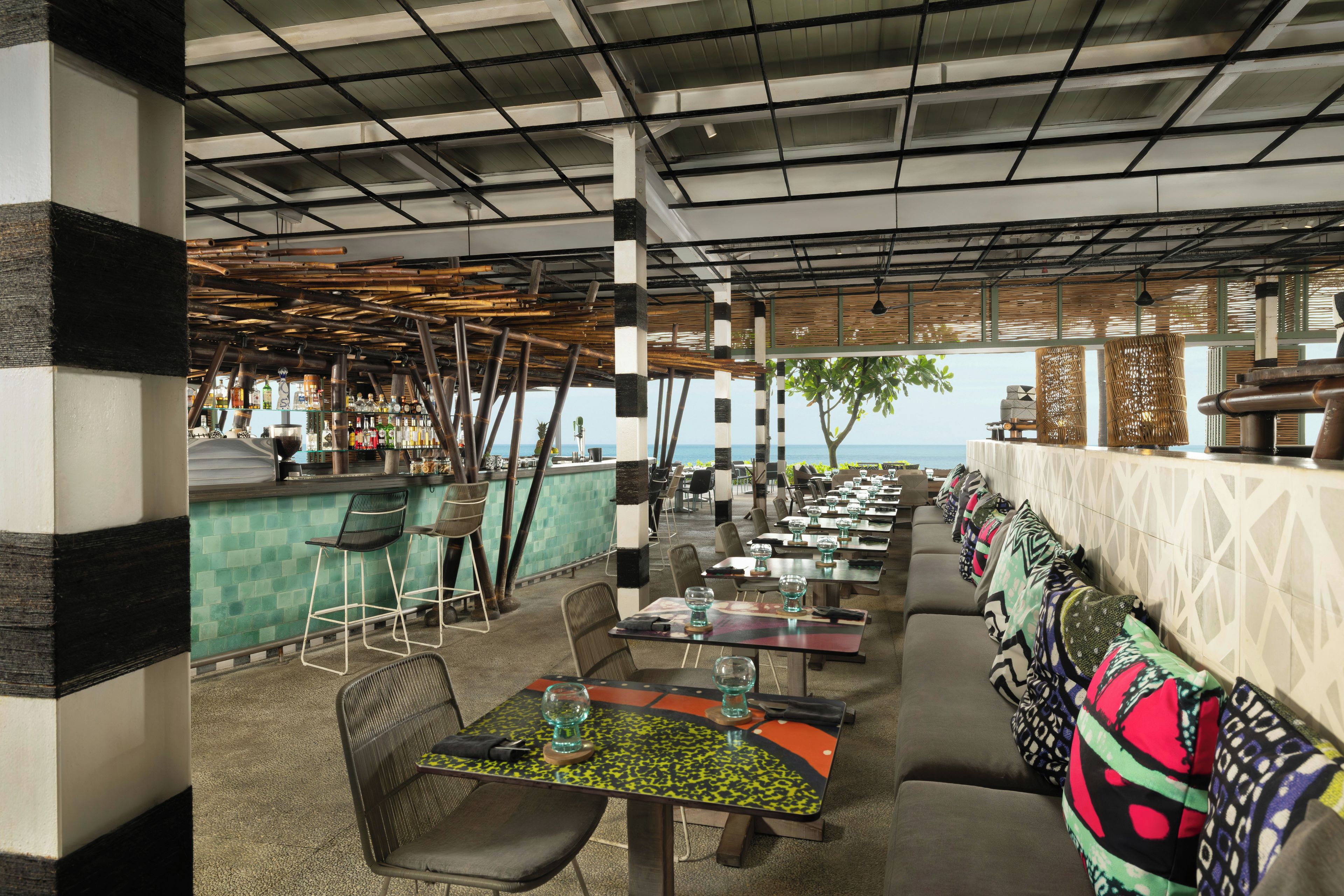 Un restaurante al aire libre con vista al mar, que cuenta con estructuras de techo de bambú, una zona de bar y arreglos de asientos.