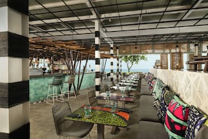 An outdoor restaurant with a view of the sea, featuring bamboo ceiling structures, a bar area, and seating arrangements.
