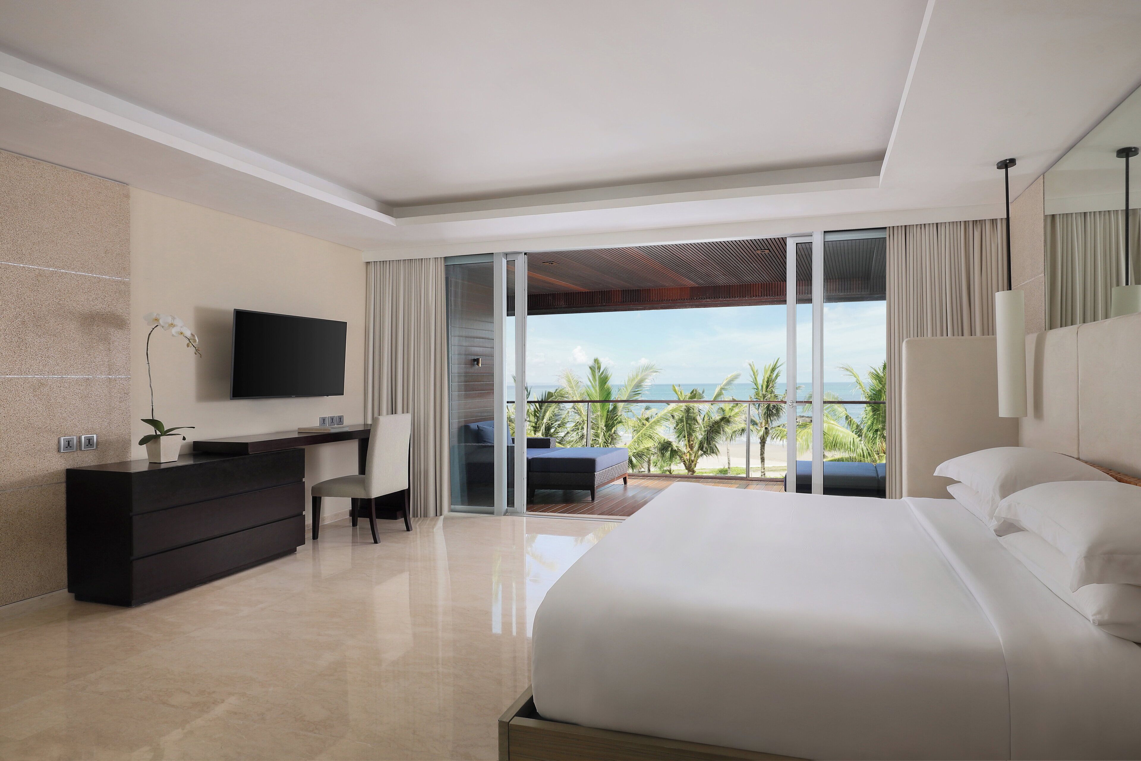 A modern hotel room with a large bed, a desk, a TV, and a balcony with a view of palm trees.