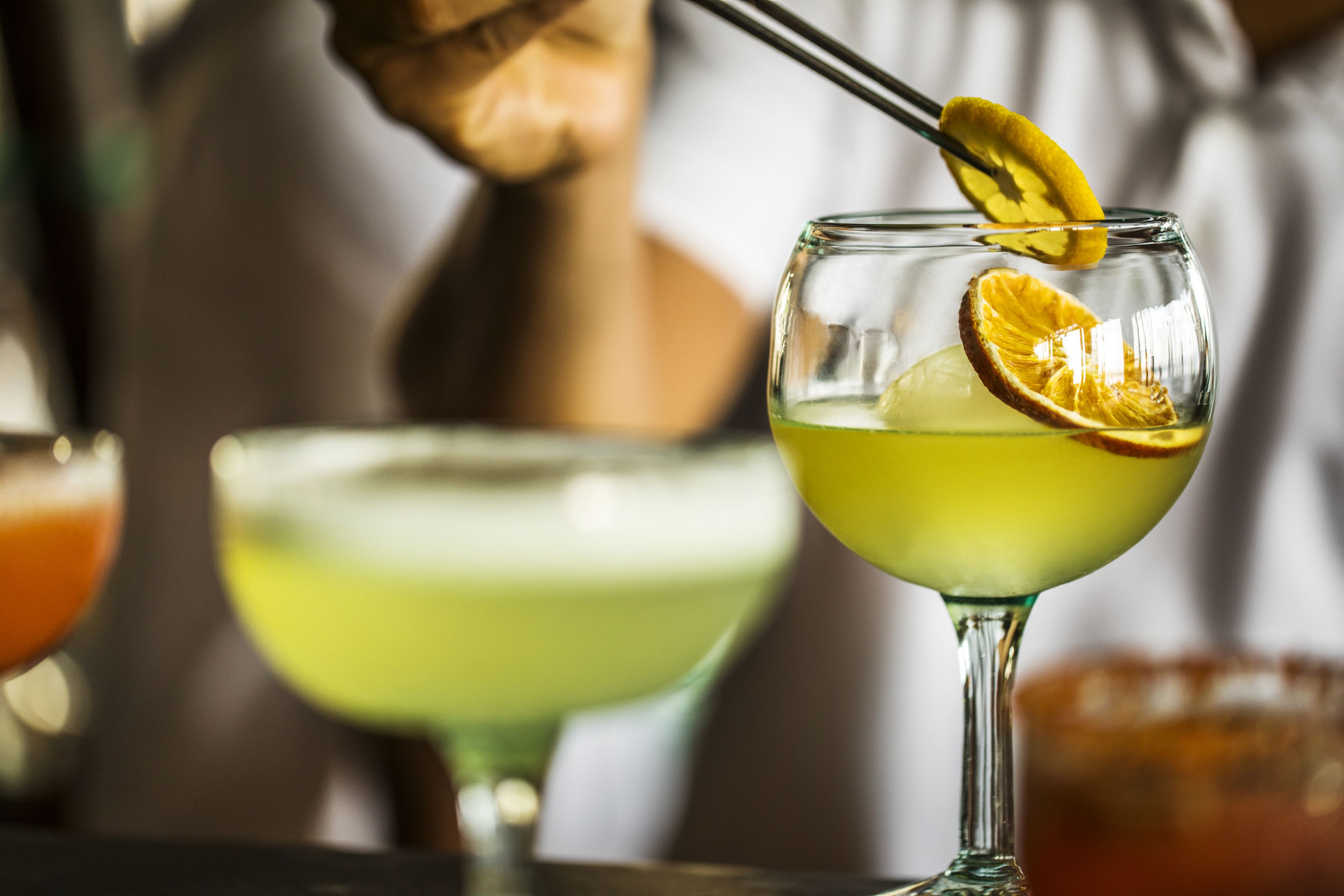 A bar setting with a cocktail glass garnished with a lemon slice and an orange slice, and another glass with a yellowish drink in the background.
