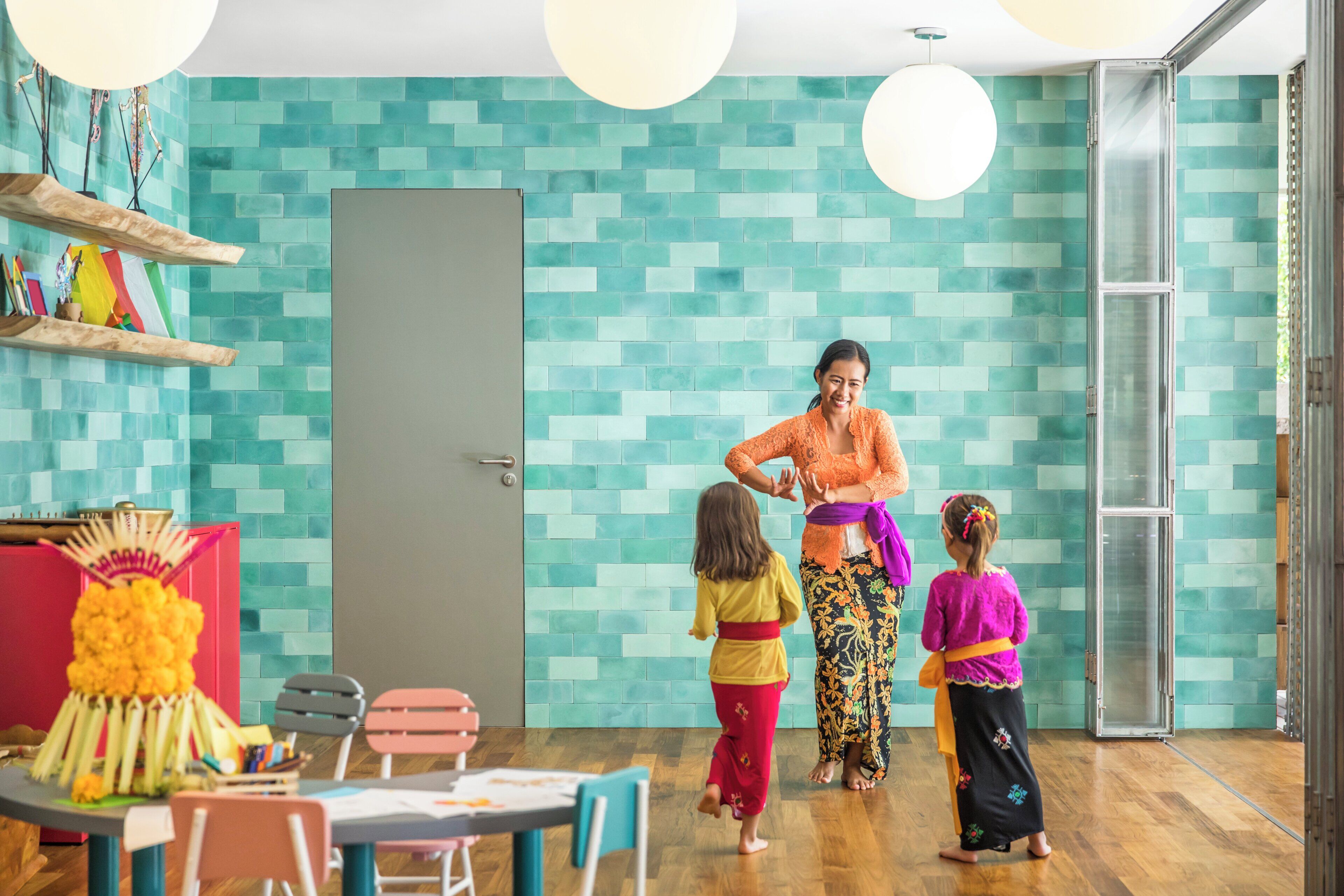 A room with teal mosaic walls, a wooden floor, and a door. There are colorful chairs and a table with a decorative item.