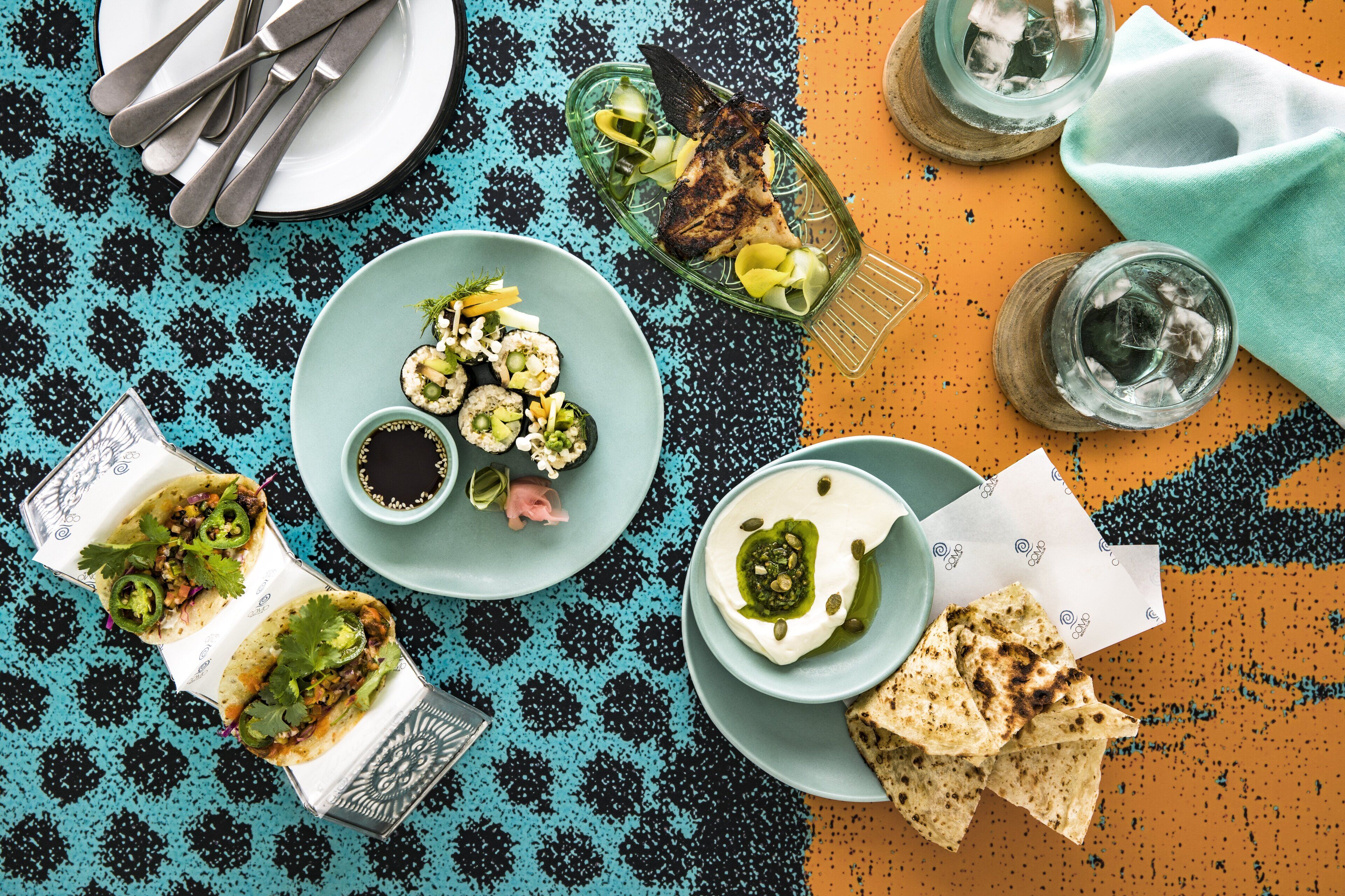 Une table où l’on trouve divers plats, notamment des tacos, des sushis et un bol de soupe, ainsi que des boissons et des couverts.