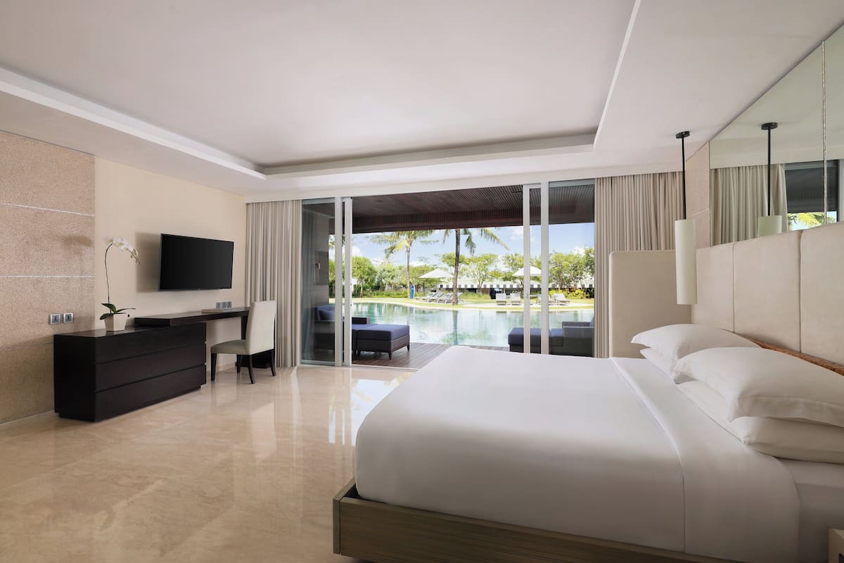 A modern hotel room with a large bed, a desk, a television, and a view of a pool and palm trees.