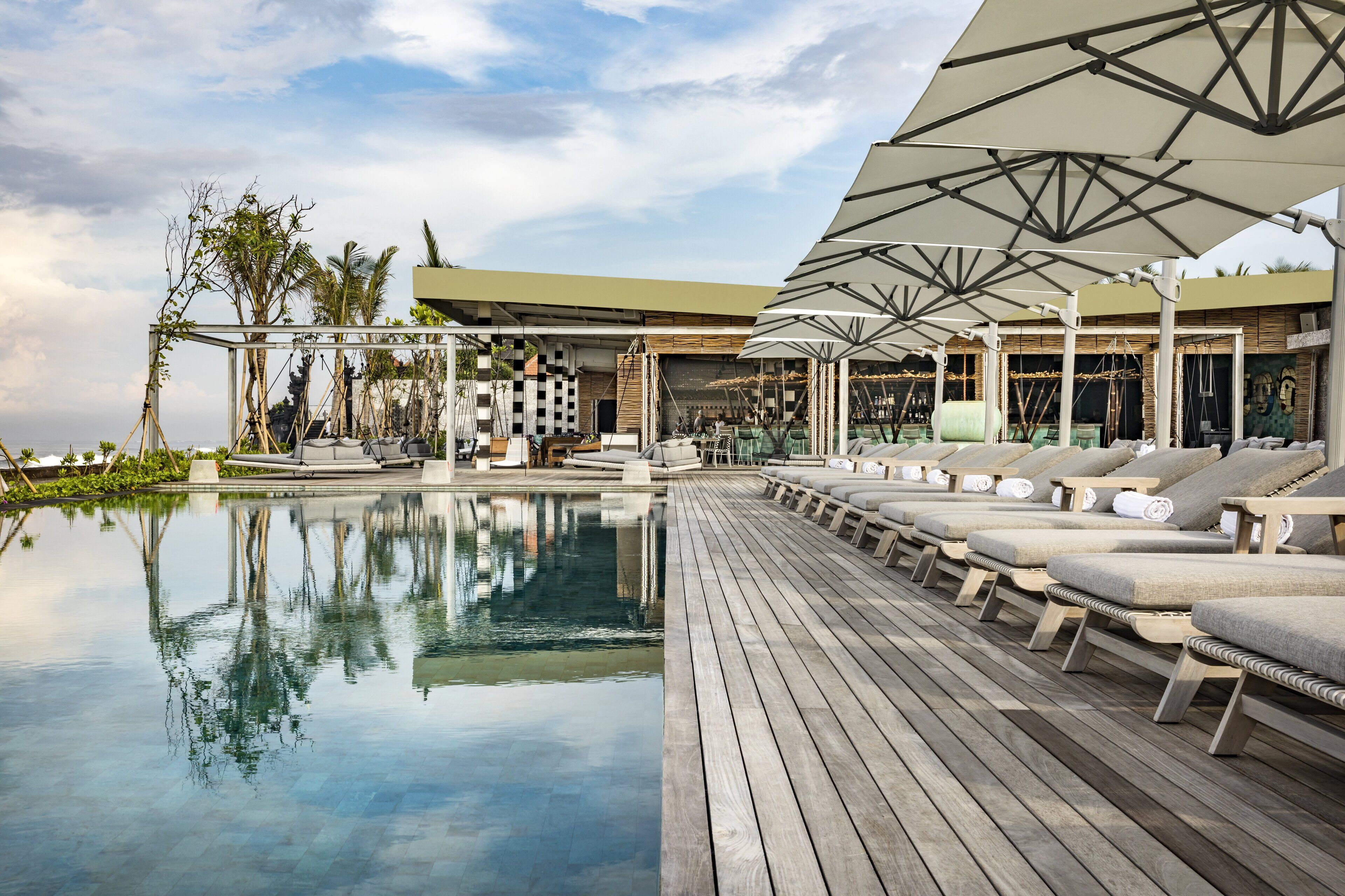 Zona de piscina con sillones de descanso y sombrillas, una terraza de madera y una piscina reflectante.