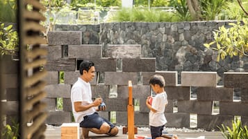 A man and a child playing a game with wooden blocks and a vertical board in an outdoor setting with a stone wall and greenery in the background.