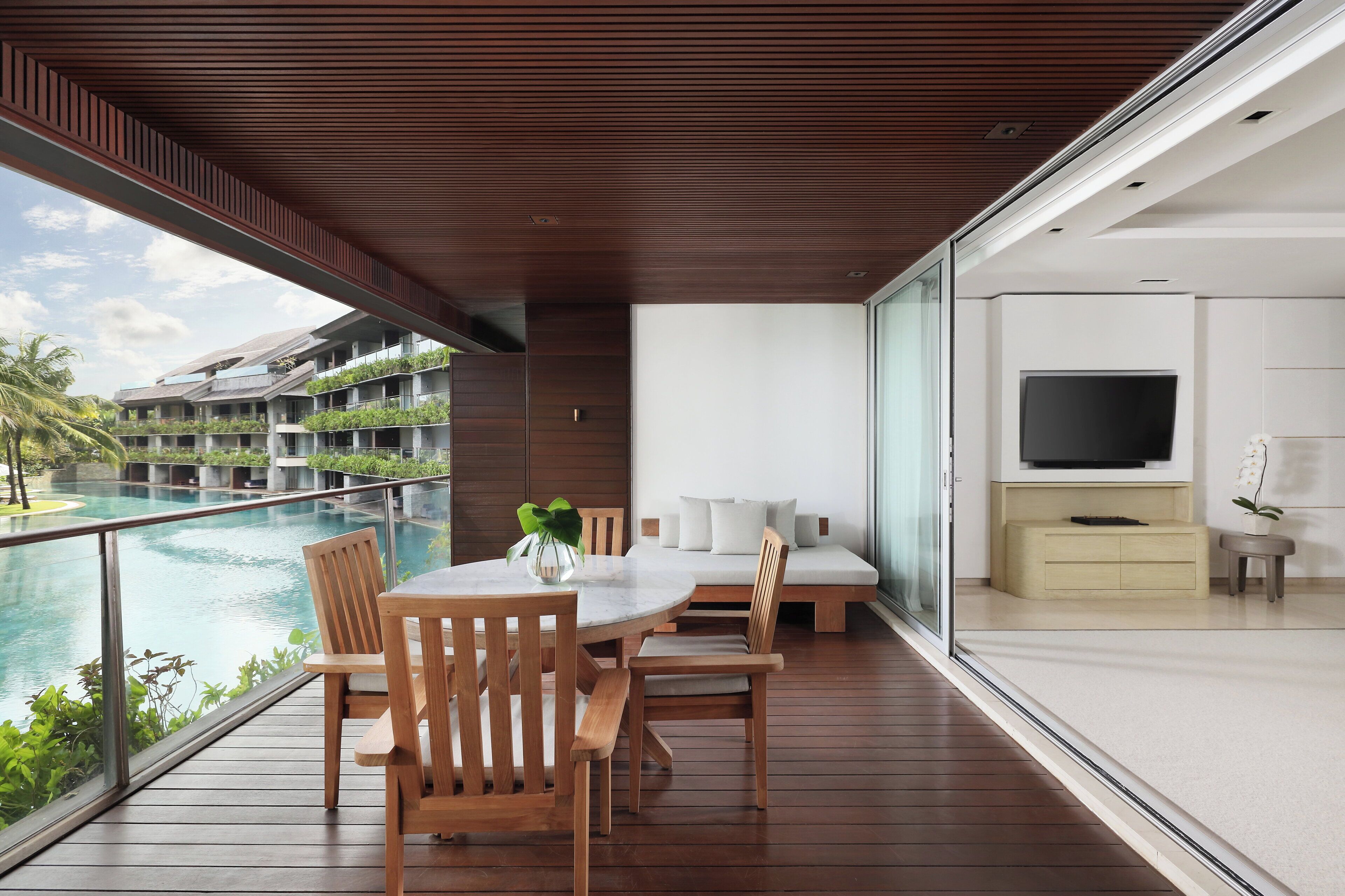 A modern balcony with a wooden table and chairs, overlooking a swimming pool and lush greenery.