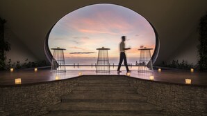 A modern interior with a large circular window overlooking a sunset, a staircase, and minimalist tables with candles.