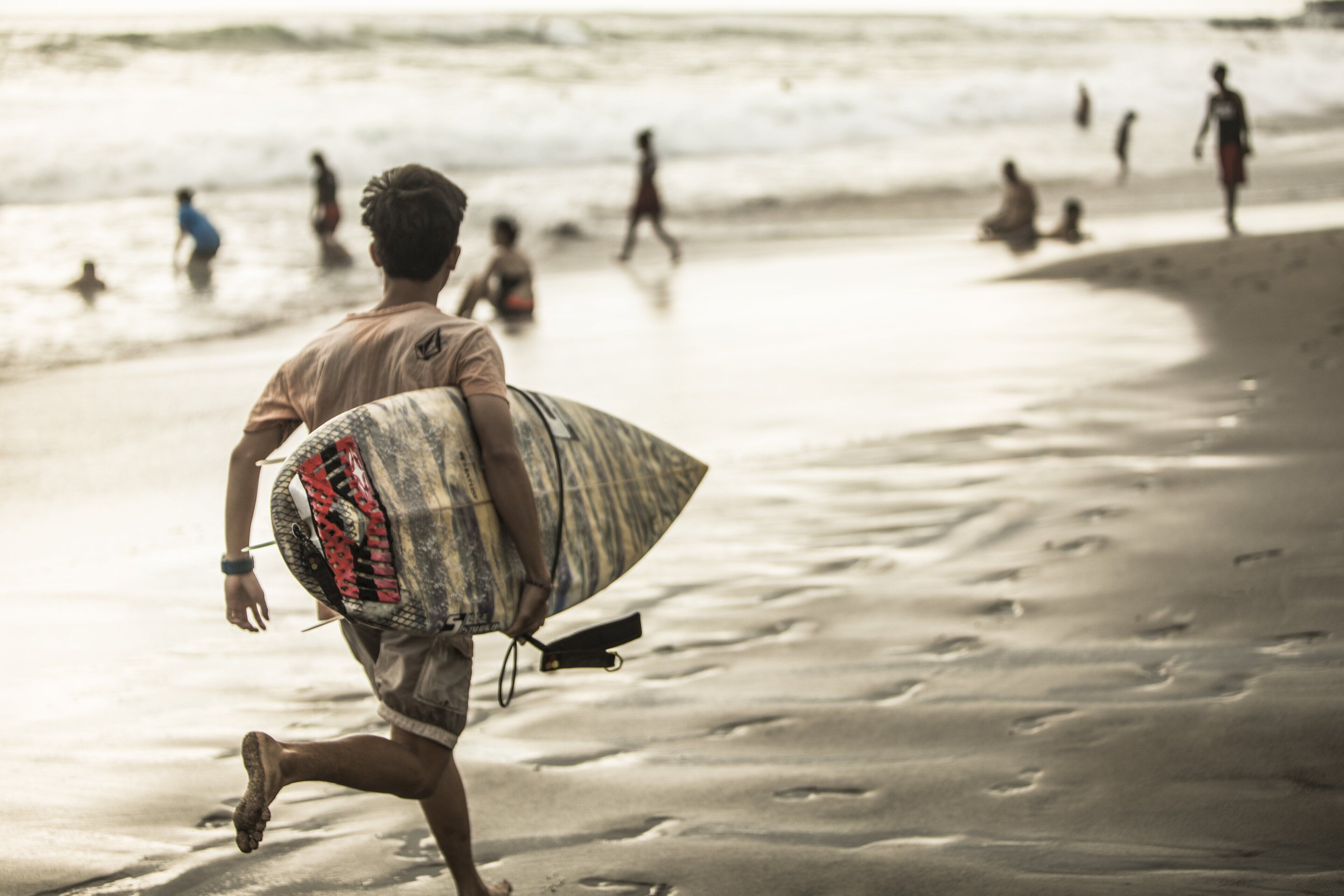 Une personne portant une planche de surf sur une plage de sable, avec des vagues en arrière-plan.