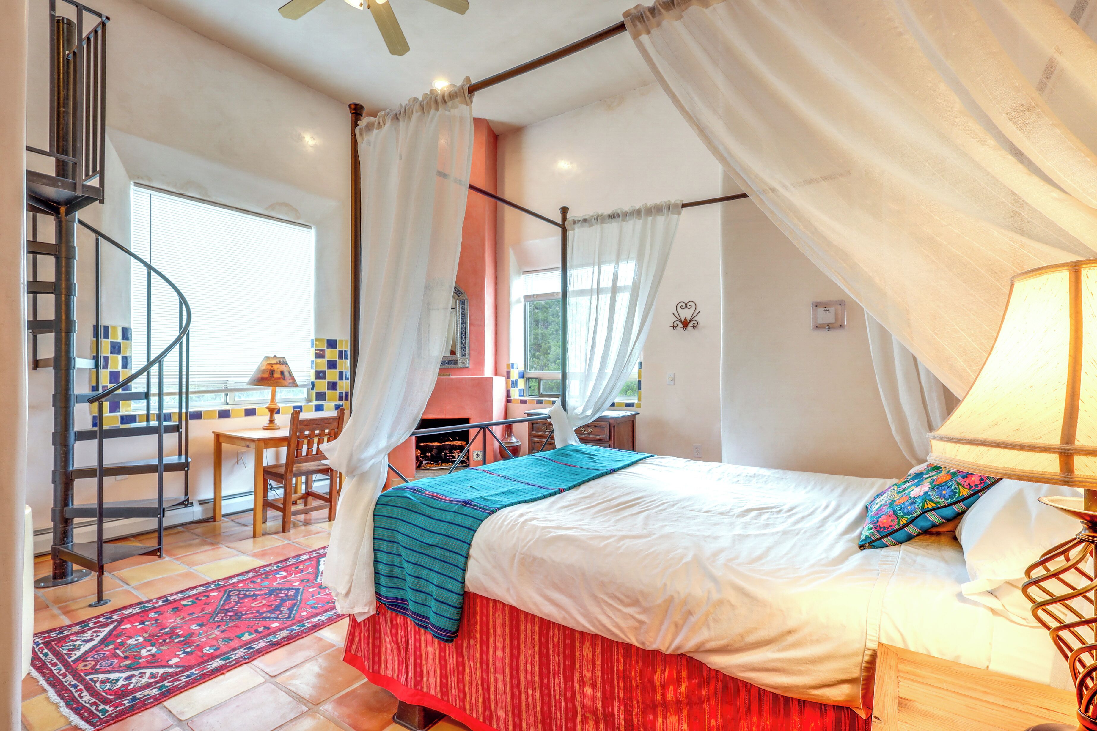 A bedroom with a bed, a red and blue striped area rug, a wooden nightstand, a lamp, and a view of a dining area with a table and chairs.
