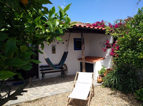 A hammock, a white chair, and a small table in a garden area with potted plants and a tiled roof. -  Tia Luísa Cottage (country house) (Redondo)