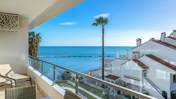 Een balkon met uitzicht op zee, rieten meubilair en witte gebouwen.