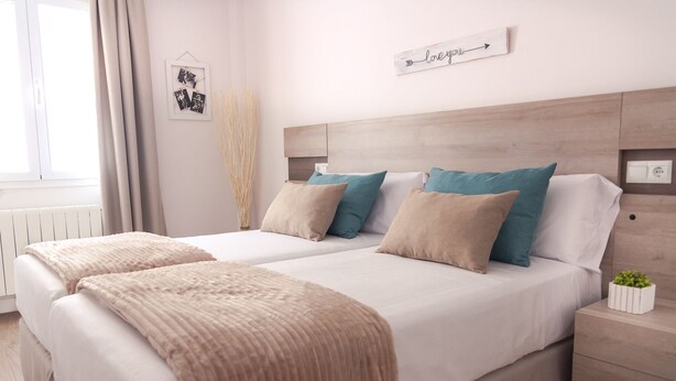 Een tweepersoonsbed met twee kussens, een nachtkastje met een plant, en een bord aan de muur.