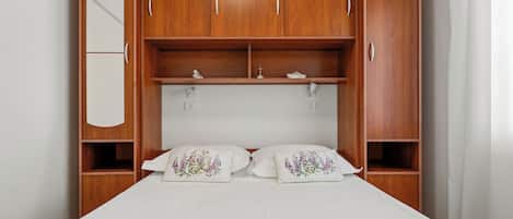 Ein Schlafzimmer mit einem Bett, zwei Kissen, einem Nachttisch, einem Spiegel und einem Holzschrank mit mehreren Fächern.