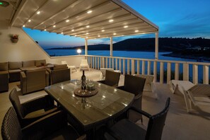 An outdoor seating area with a table set for dining, overlooking a body of water at dusk.