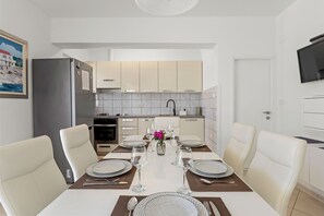 A modern kitchen with white cabinets, a built-in oven, and a dining area set for four with white chairs and a table set with plates, glasses, and cutlery.