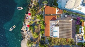 Vista aerea di una proprietà costiera con spiaggia, una barca ormeggiata vicino alla riva e una casa con tetto in tegole e area salotto esterna.