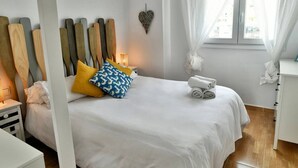 A neatly made bed with white bedding and a few decorative pillows, a bedside table with a lamp, and a window with sheer curtains.