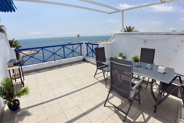 Un patio exterior con mesa y sillas, vista al mar y barandilla azul.