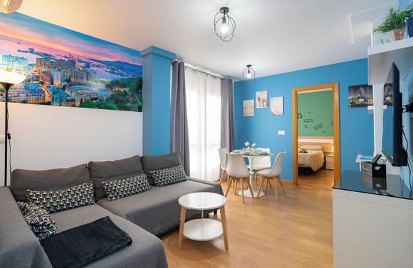 A modern living room with a sofa, dining table, and chairs. There is a painting on the wall, a ceiling light, and a bed in the adjacent room.