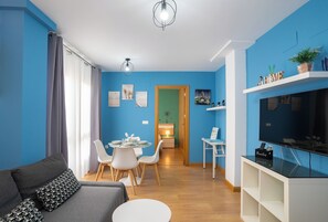 A modern living room with a blue wall, a grey sofa, a dining area with a table and chairs, and a TV on a white stand.