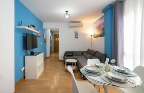 A modern living room with a flat-screen TV, a sofa, a dining table set for four, and a hallway leading to a bedroom.