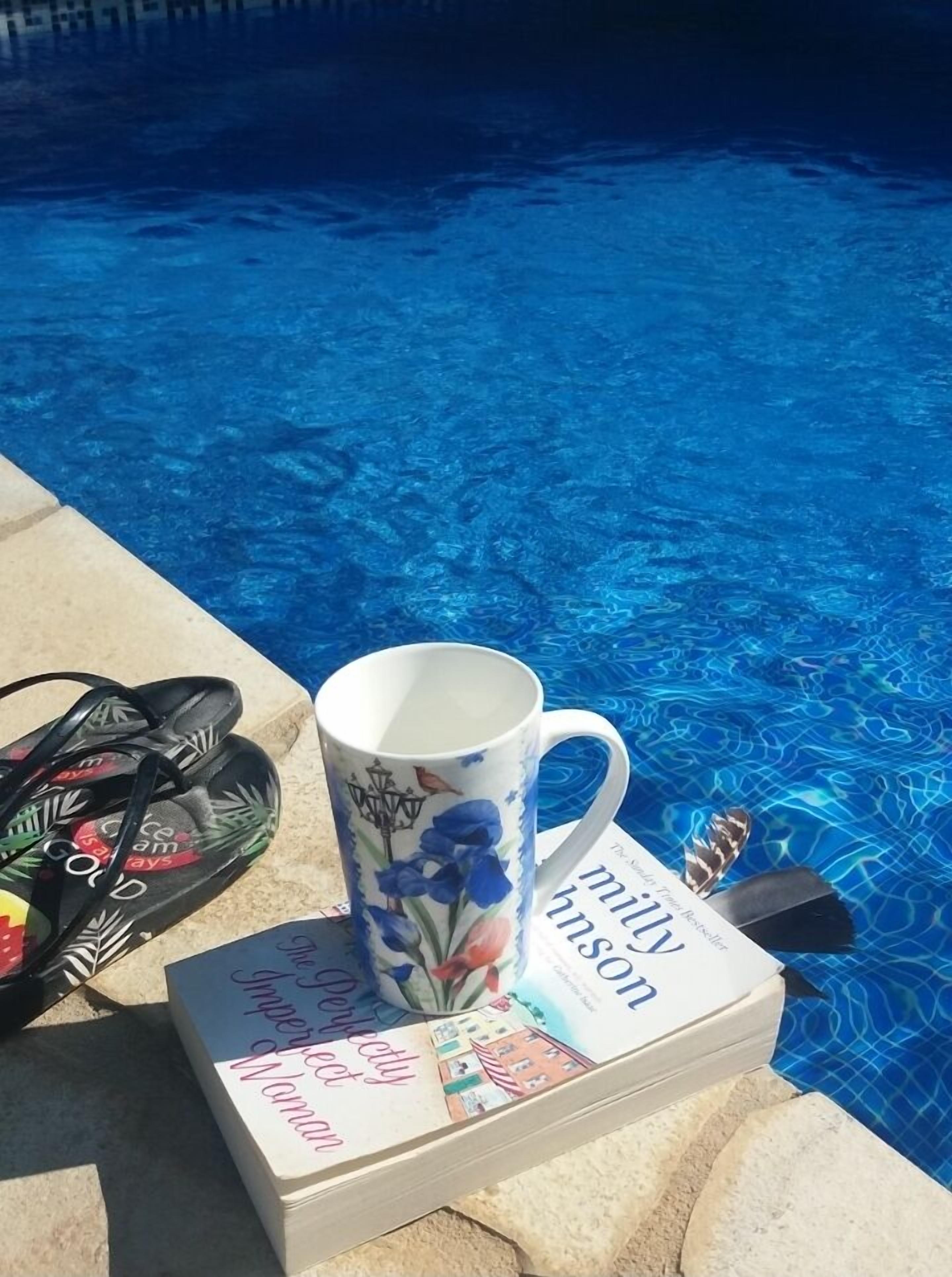 A poolside area with a floral mug, a book titled 'Milly Knox', and a pair of sandals.