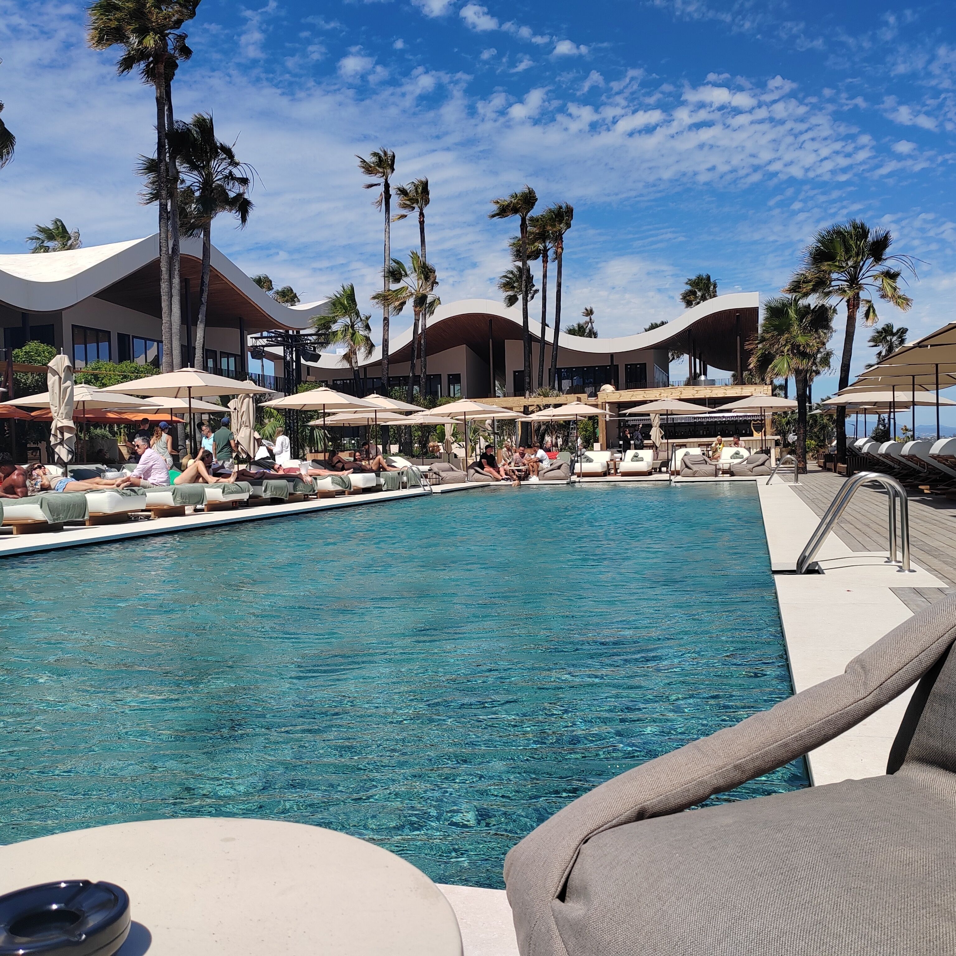 A pool area with lounge chairs and umbrellas, surrounded by palm trees and modern architecture.