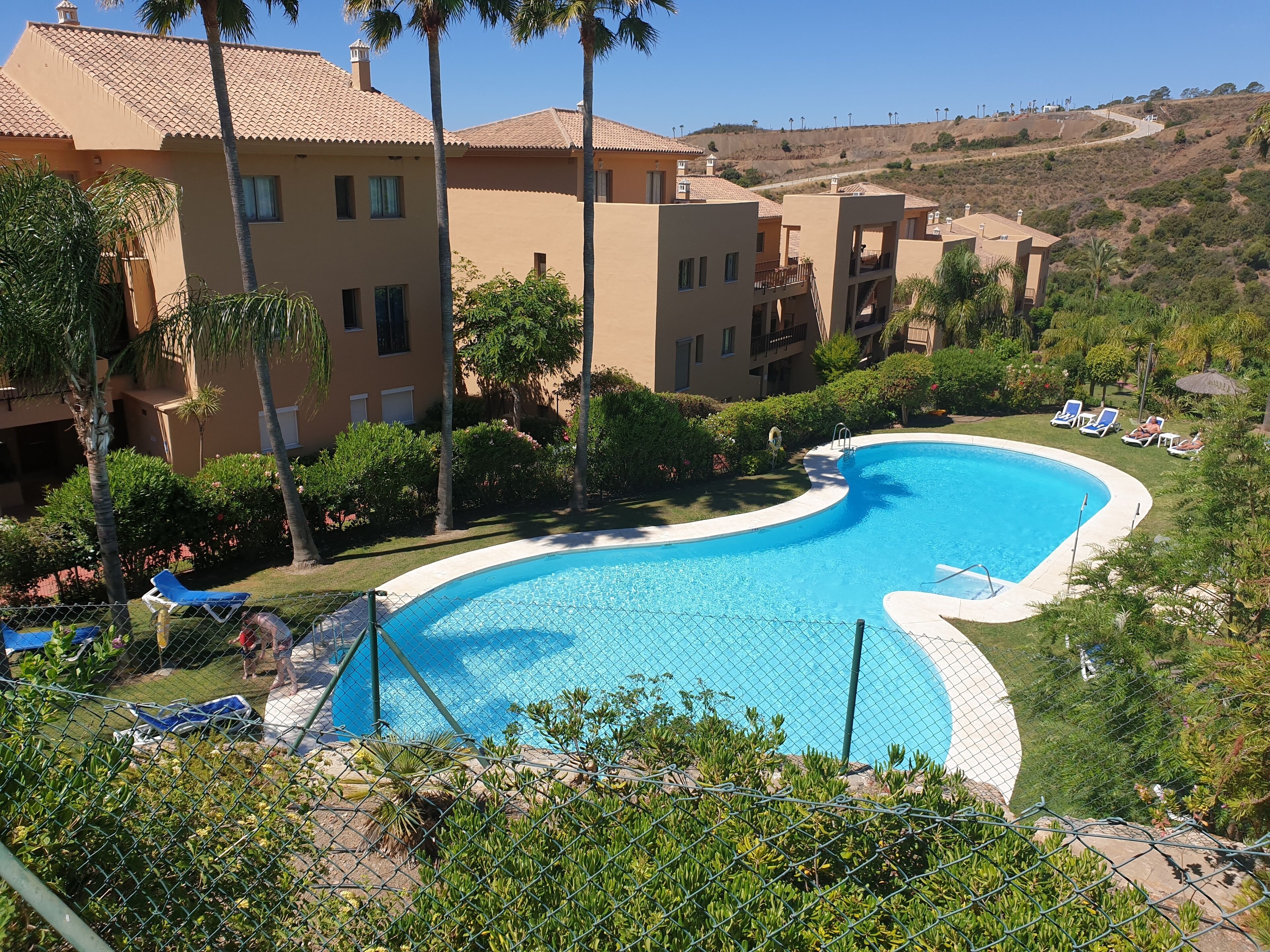 Una piscina circondata da una recinzione, con vista su un pendio e edifici sullo sfondo.
