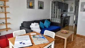 Un espacio habitable compacto con un sofá gris, cojines azules, una mesa de comedor de madera y una zona de cocina con microondas y estufa.