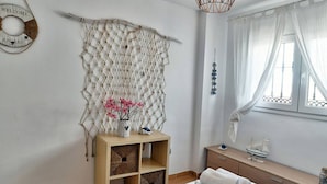 Un dormitorio con cama, una mesita de noche con un jarrón de flores, un adorno en la pared y una ventana con cortinas.