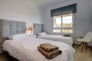 A hotel room with two beds, a window, a chair, and a lamp.